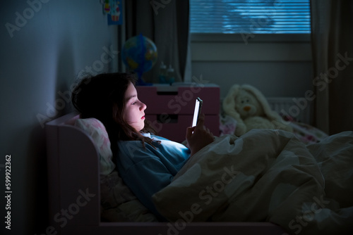 A child using smart phone lying in bed late at night, playing games, watching videos online, scrolling screen. Children's screen addiction. Child's room at night. 
