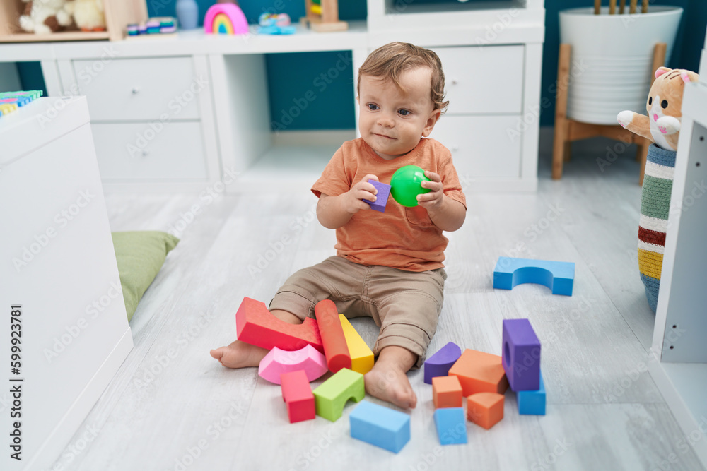 Adorable blond toddler playing with ball and geometry blocks sitting on floor at kindergarten Adorable blond toddler playing with ball and geometry blocks sitting on floor at kindergarten