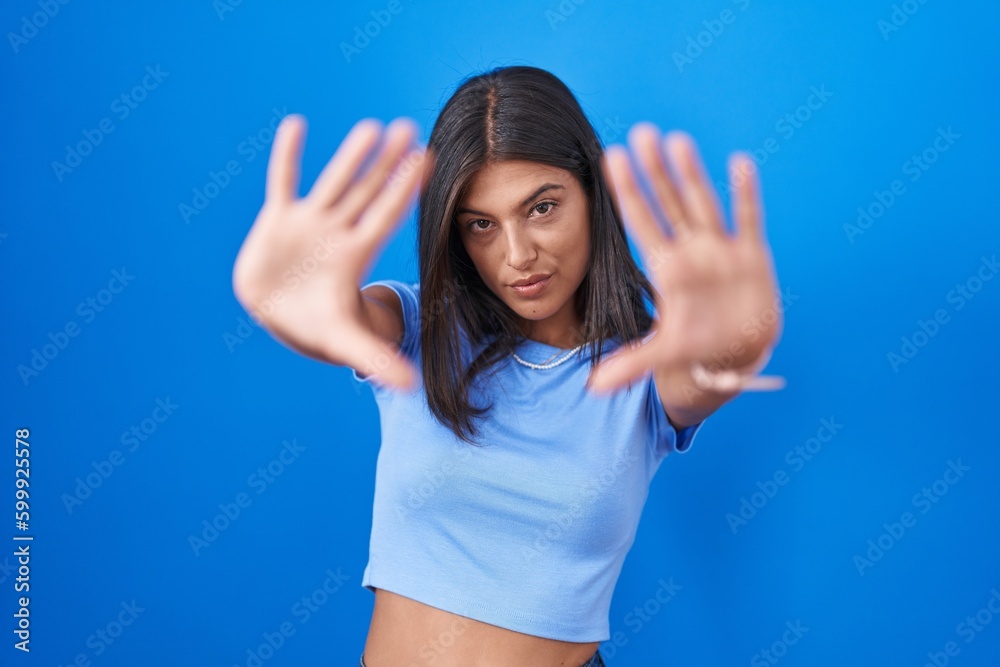 Brunette young woman standing over blue background doing frame using hands palms and fingers, camera perspective Brunette young woman standing over blue background doing frame using hands palms and fingers, camera perspective
