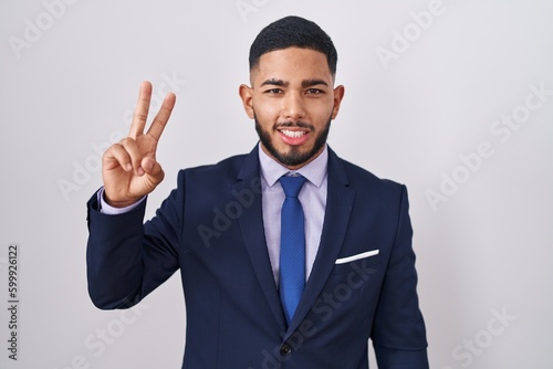 Young hispanic man wearing business suit and tie smiling looking to the camera showing fingers doing victory sign. number two.