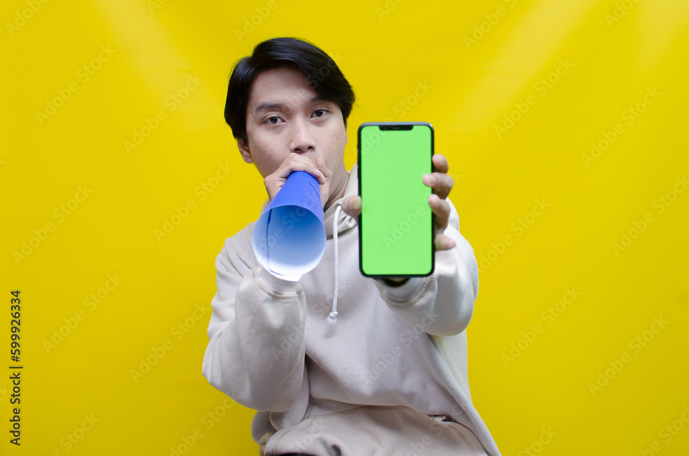 Ecstatic Asian man in beige hoodie holding megaphone from paper showing green phone screen. man holding megaphone from paper giving announcement and promotion isolated over yellow background