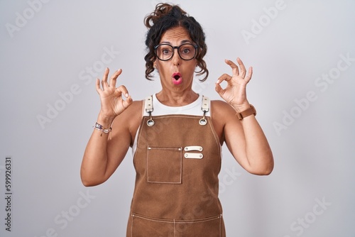 Obraz na płótnie Middle age woman wearing apron over white background looking surprised and shocked doing ok approval symbol with fingers