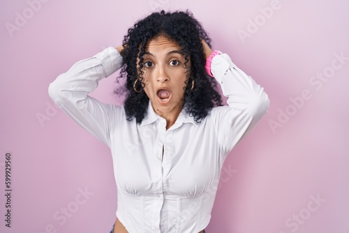 Hispanic woman with curly hair standing over pink background crazy and scared with hands on head, afraid and surprised of shock with open mouth