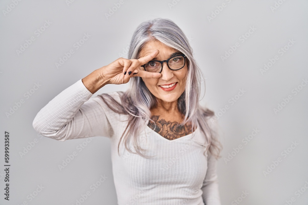 Middle age woman with grey hair standing over white background doing peace symbol with fingers over face, smiling cheerful showing victory Middle age woman with grey hair standing over white background doing peace symbol with fingers over face, smiling cheerful showing victory