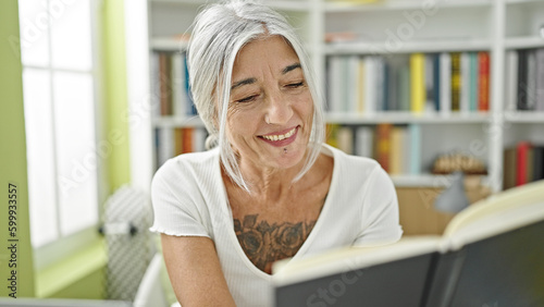 Middle age grey-haired woman student reading book smiling at library university