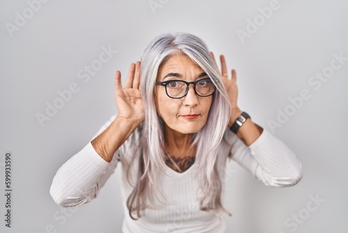 Middle age woman with grey hair standing over white background trying to hear both hands on ear gesture, curious for gossip. hearing problem, deaf