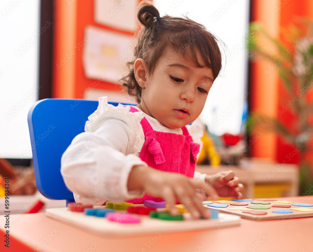 Adorable hispanic girl playing with maths puzzle game sitting on table at kindergarten