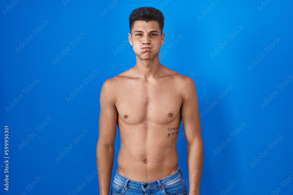 Young hispanic man standing shirtless over blue background puffing cheeks with funny face. mouth inflated with air, crazy expression.