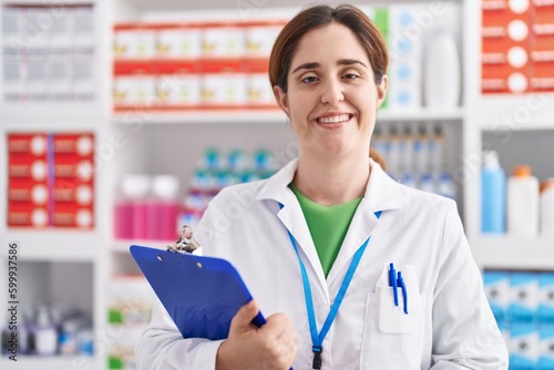 Young woman pharmacist smiling confident holding clipboard at pharmacy