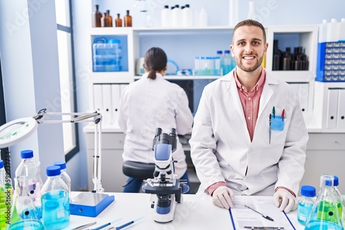 Young man working at scientist laboratory looking positive and happy standing and smiling with a confident smile showing teeth