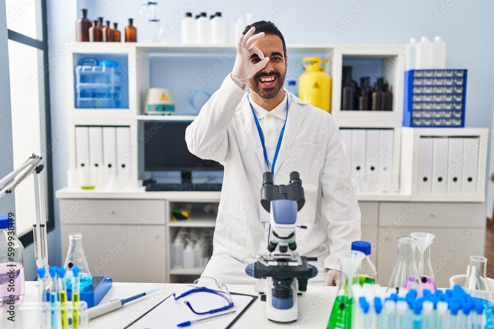 Young hispanic man with beard working at scientist laboratory doing ok gesture with hand smiling, eye looking through fingers with happy face.