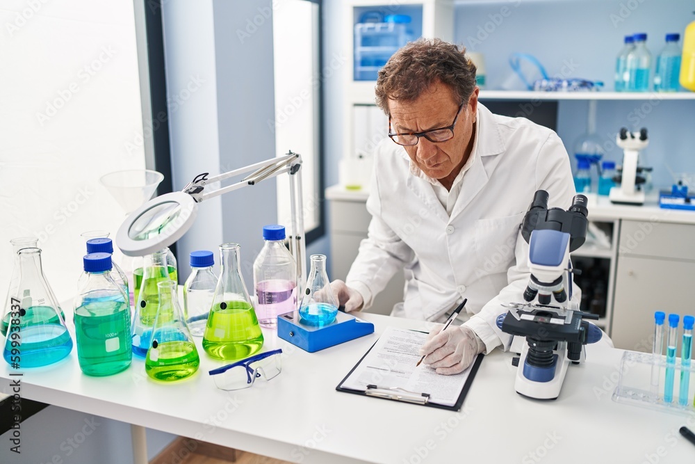 Middle age man wearing scientist uniform measuring test tube writing on clipboard at laboratory Middle age man wearing scientist uniform measuring test tube writing on clipboard at laboratory