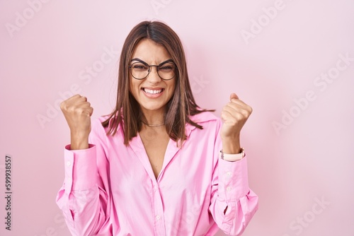 Young hispanic woman wearing glasses standing over pink background very happy and excited doing winner gesture with arms raised  smiling and screaming for success. celebration concept.