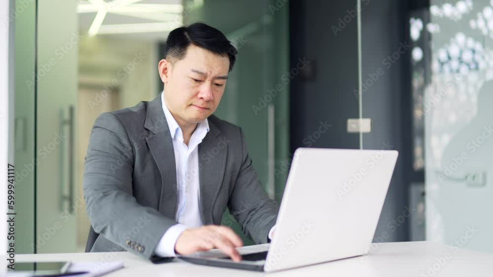 Frustrated asian man having problems with software or service on laptop while sitting at workplace in modern office. A worried businessman in a formal suit complains about a bad internet connection Frustrated asian man having problems with software or service on laptop while sitting at workplace in modern office. A worried businessman in a formal suit complains about a bad internet connection