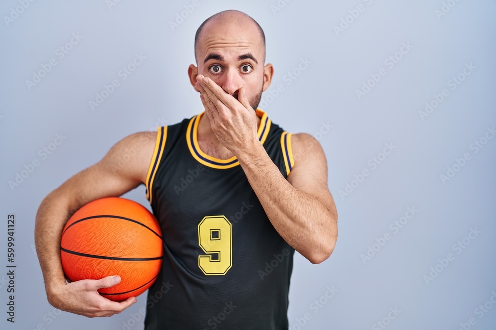 Young bald man with beard wearing basketball uniform holding ball shocked covering mouth with hands for mistake. secret concept. Young bald man with beard wearing basketball uniform holding ball shocked covering mouth with hands for mistake. secret concept.