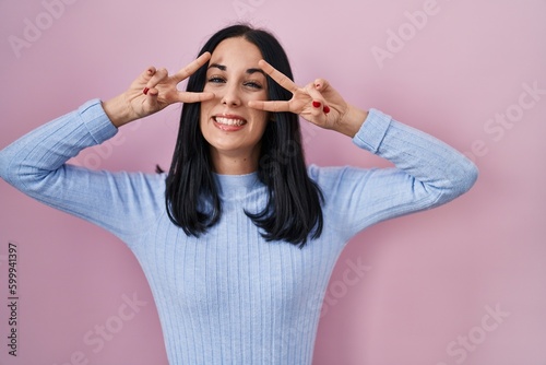 Hispanic woman standing over pink background doing peace symbol with fingers over face, smiling cheerful showing victory