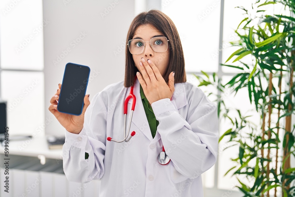 Young hispanic doctor woman showing smartphone screen covering mouth with hand, shocked and afraid for mistake. surprised expression Young hispanic doctor woman showing smartphone screen covering mouth with hand, shocked and afraid for mistake. surprised expression