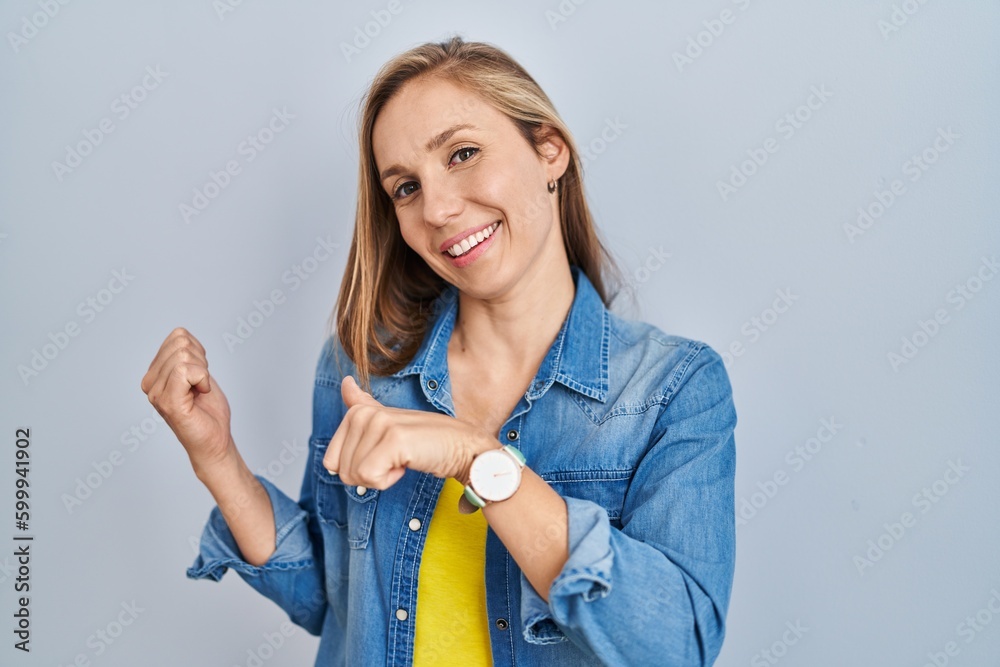 Young blonde woman standing over blue background pointing to the back behind with hand and thumbs up, smiling confident Young blonde woman standing over blue background pointing to the back behind with hand and thumbs up, smiling confident
