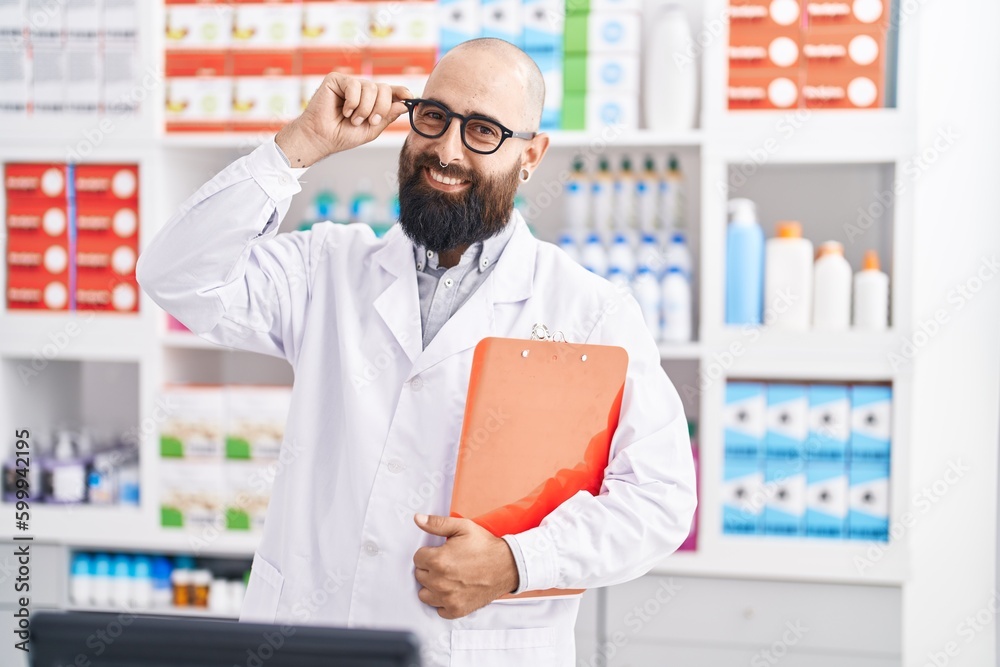Young bald man pharmacist smiling confident holding clipboard at pharmacy
