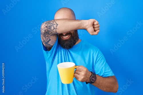 Young hispanic man with beard and tattoos drinking a cup of coffee smiling cheerful playing peek a boo with hands showing face. surprised and exited