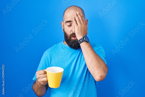 Young hispanic man with beard and tattoos drinking a cup of coffee yawning tired covering half face, eye and mouth with hand. face hurts in pain. © Krakenimages.com