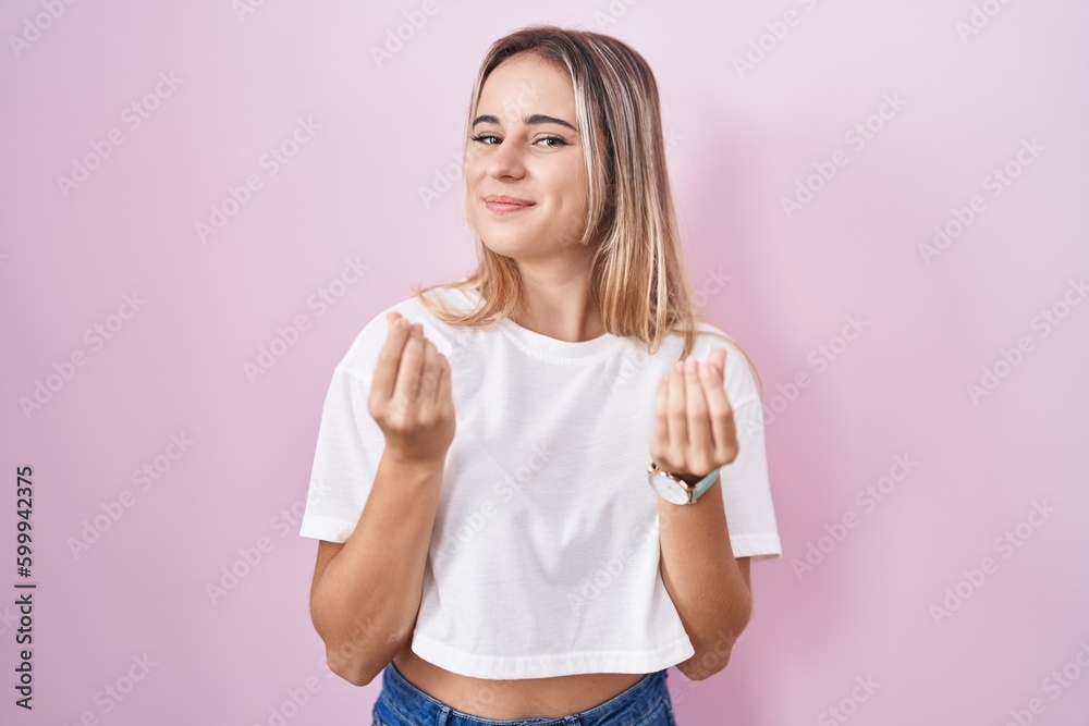 Young blonde woman standing over pink background doing money gesture with hands, asking for salary payment, millionaire business