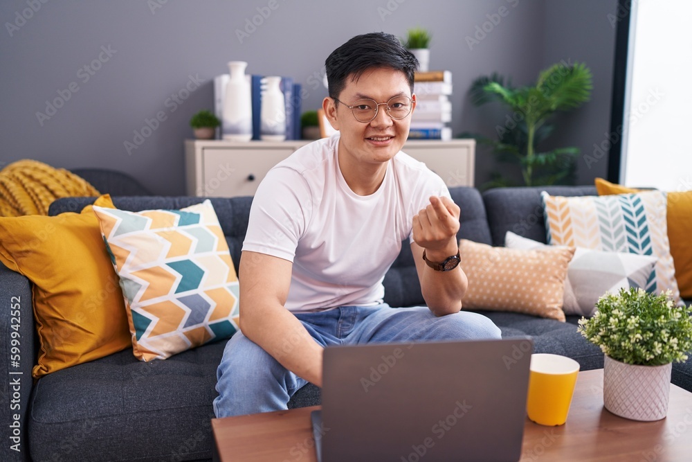 Young asian man using laptop at home sitting on the sofa beckoning come here gesture with hand inviting welcoming happy and smiling Young asian man using laptop at home sitting on the sofa beckoning come here gesture with hand inviting welcoming happy and smiling