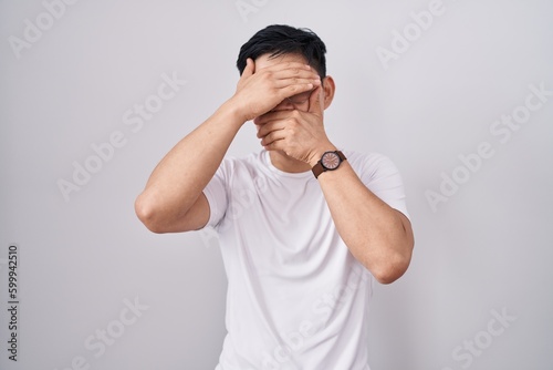 Young asian man standing over white background covering eyes and mouth with hands, surprised and shocked. hiding emotion