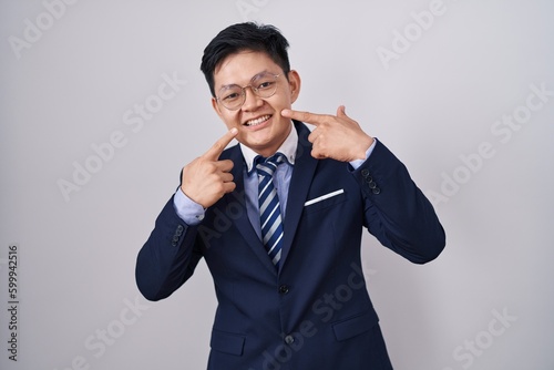 Young asian man wearing business suit and tie smiling cheerful showing and pointing with fingers teeth and mouth. dental health concept.