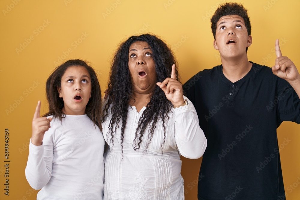 Family of mother, daughter and son standing over yellow background amazed and surprised looking up and pointing with fingers and raised arms. Family of mother, daughter and son standing over yellow background amazed and surprised looking up and pointing with fingers and raised arms.