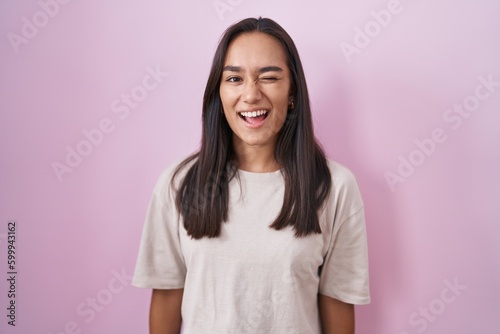 Young hispanic woman standing over pink background winking looking at the camera with sexy expression  cheerful and happy face.