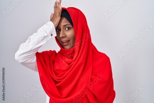 Young arab woman wearing traditional islamic hijab scarf surprised with hand on head for mistake, remember error. forgot, bad memory concept.