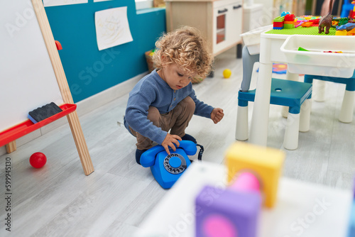Adorable blond toddler playing with telephone toy sitting on floor at kindergarten