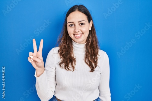 Young hispanic woman standing over blue background showing and pointing up with fingers number two while smiling confident and happy.
