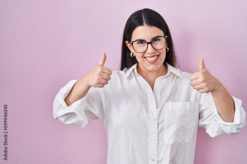 Young brunette woman standing over pink background success sign doing positive gesture with hand, thumbs up smiling and happy. cheerful expression and winner gesture. Young brunette woman standing over pink background success sign doing positive gesture with hand, thumbs up smiling and happy. cheerful expression and winner gesture.