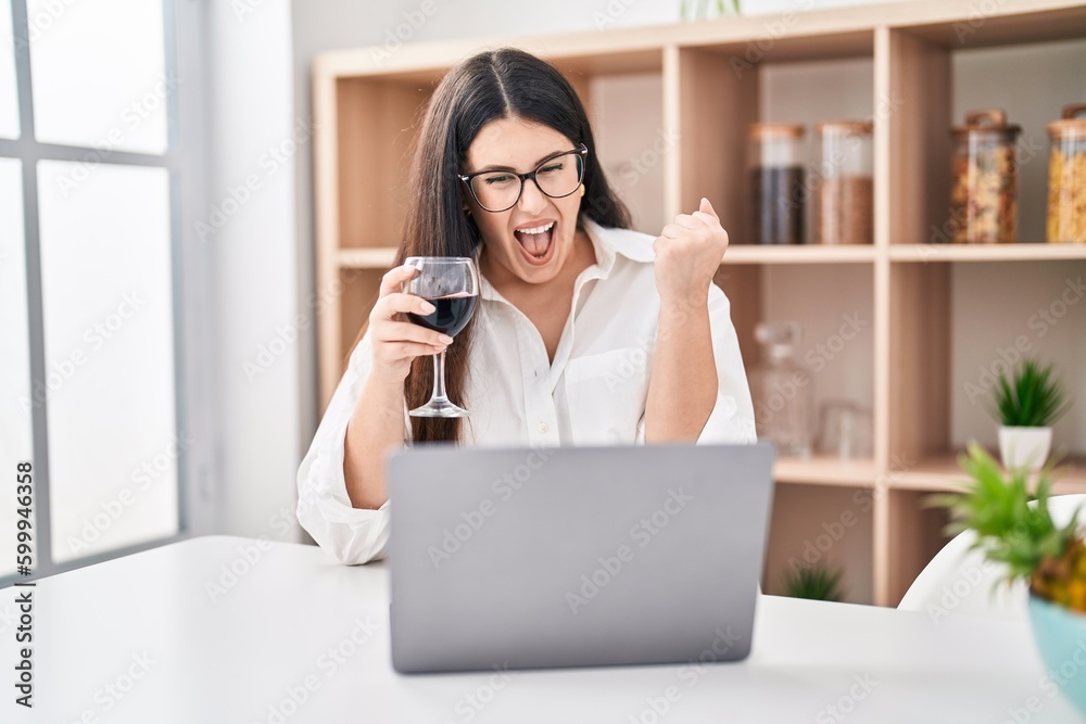 Young brunette woman doing video call with laptop drinking a glass of wine screaming proud, celebrating victory and success very excited with raised arm Young brunette woman doing video call with laptop drinking a glass of wine screaming proud, celebrating victory and success very excited with raised arm