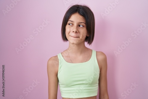 Young girl standing over pink background smiling looking to the side and staring away thinking.