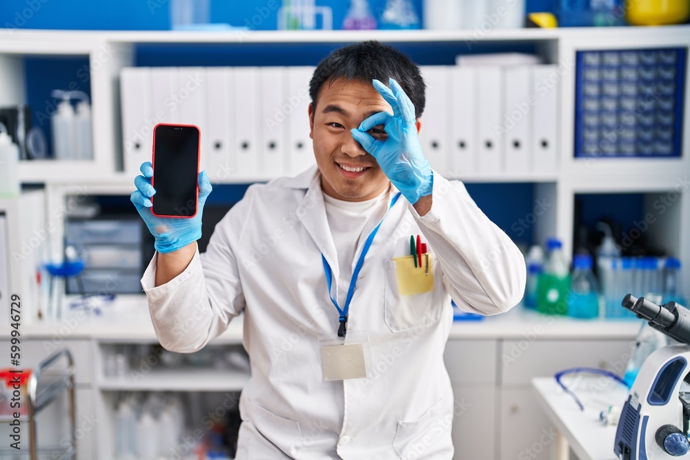 Young chinese man working at scientist laboratory holding smartphone smiling happy doing ok sign with hand on eye looking through fingers