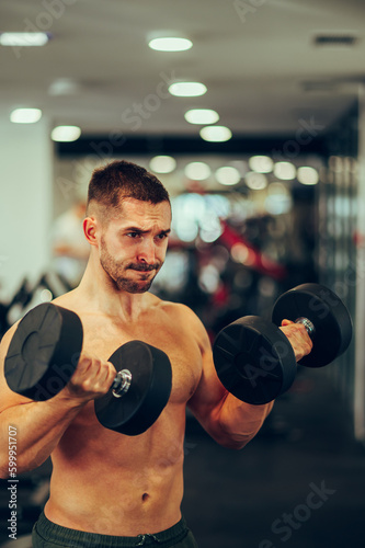 A focused fit sportsman without a shirt is lifting heavy dumbbells in a gym and doing exercises for the biceps.