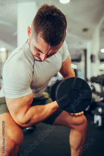 Close up of a focused strong man flexing and pumping muscles with a dumbbell while sitting in a gym.