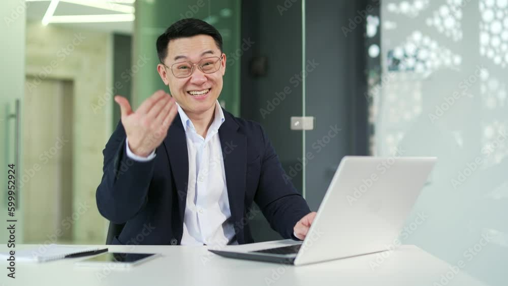 Smiling asian man pointing finger at viewer and calling to him with hand gesture looking at camera while sitting at desk at workplace in office. Happy boss, owner in a formal suit calls to his company Smiling asian man pointing finger at viewer and calling to him with hand gesture looking at camera while sitting at desk at workplace in office. Happy boss, owner in a formal suit calls to his company