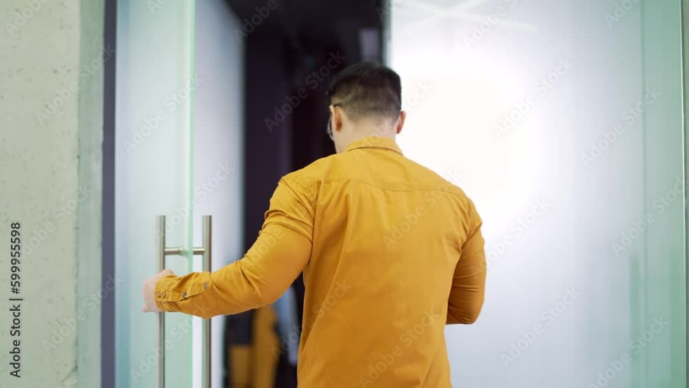 Rear view. A man in a shirt opens a glass door in a modern building and enters the room. A male businessman or employee goes to the office to the workplace holding a smartphone in his hands Rear view. A man in a shirt opens a glass door in a modern building and enters the room. A male businessman or employee goes to the office to the workplace holding a smartphone in his hands