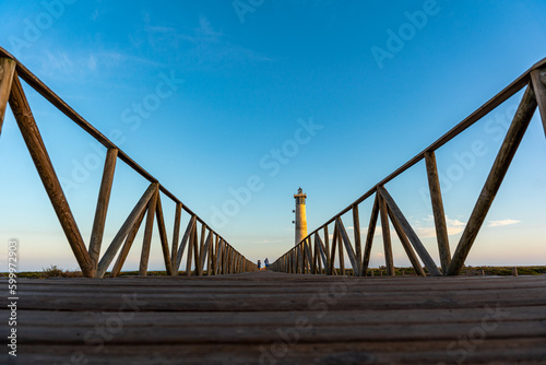 puente de madera con faro de fondo y dos personas caminando en el atardecer
