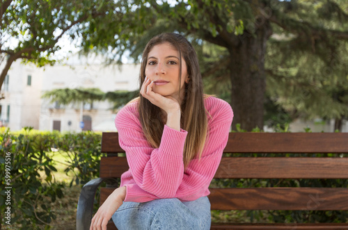 young beautiful pensive woman with long light brown hair sitting fuchsia sweater and park background with hand supporting head under chin