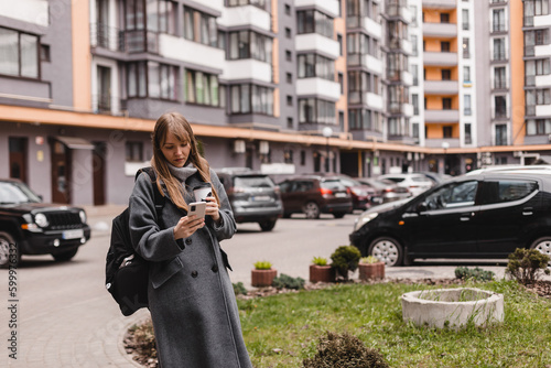 Young blonde woman with bang in casual wear smiling while using smartphone and holding takeaway coffee outdoors. Blonde woman wear grey coat and sweater, black backpack, typing message, read news.