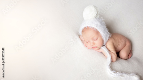 a newborn baby in a white hat with a pom-pom sleeps on a white bedspread over white background with copy space. beautiful sleeping baby. white banner