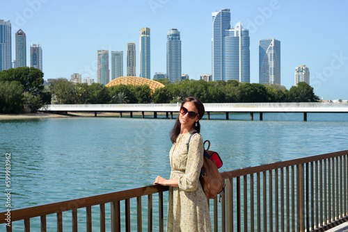 The skyline of Sharjah is visible from the waterfront - a woman tourist with a backpack enjoys the view of the water and skyscrapers. Cityscape of Sharjah UAE.