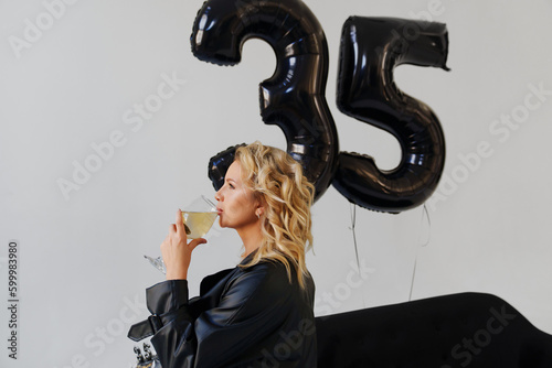 a blonde in a black with champagne on the couch. balloons of numbers thirty-five