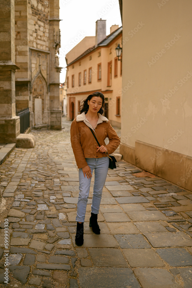 Young Asian model in fashionable clothing posing on a historic street in a European city. Beautiful cityscape and landmarks in the background. Ideal for travel, tourism, and fashion-related themes.