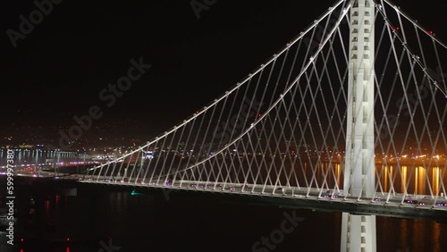 Aerial Panning Shot Of Illuminated Oakland Bay Bridge On Sea At Night, Drone Flying Near Famous Landmark Against Sky - San Francisco, California photo